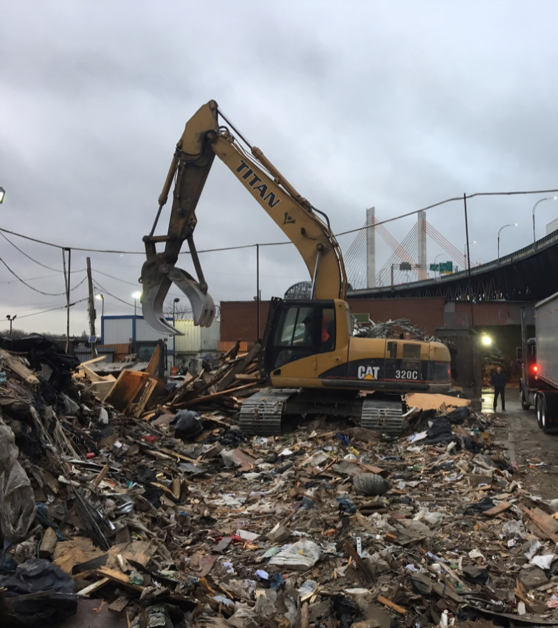 Excavator with grapple handling construction and demolition debris at a recycling facility.