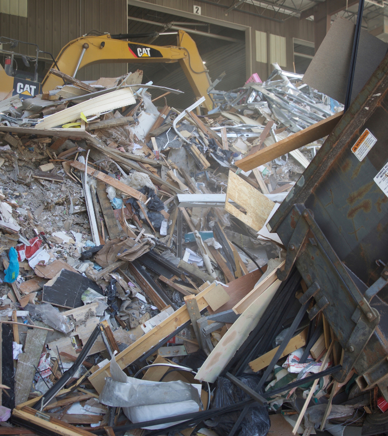 Mixed construction and demolition debris at a recycling facility with an excavator in the background.